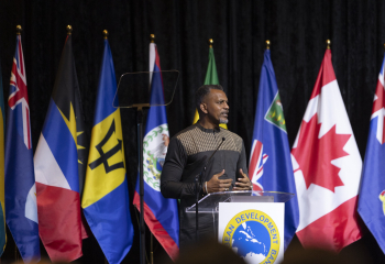 President of the Caribbean Development Bank delivers remarks from a podium on stage, with multiple national flags displayed behind him.