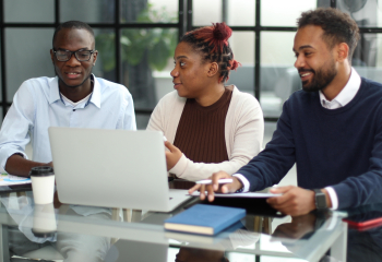 Three youth sitting in a boardroom in front of a laptop having a conversation. 