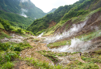Steaming geothermal vents in a lush green mountain valley, with a hiker walking along a rocky trail in the foreground