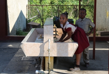 A schoolgirl in red and white school uniform is washing hands at an outside lavatory while a schoolboy in sand uniform is waiting behind her