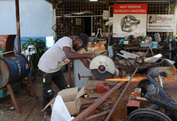 A trainee wearing a black baseball cap and trousers, a sand T-shirt, and protective goggles is cutting a piece of metal with a disc saw
