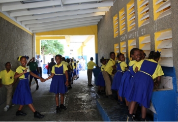 Female students, wearing yellow shirts and blue skirts, and male students, wearing yellow shirts and kaki trousers, are walking through a covered passageway with lavatories