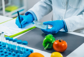 lab tech in white jacket and blue gloves with vegetables and vials on desk