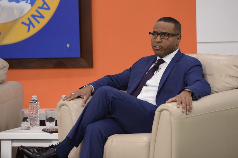 Dr. Justin Ram wearing a blue suit, white shirt and mauve tie seated on a cream soft chair