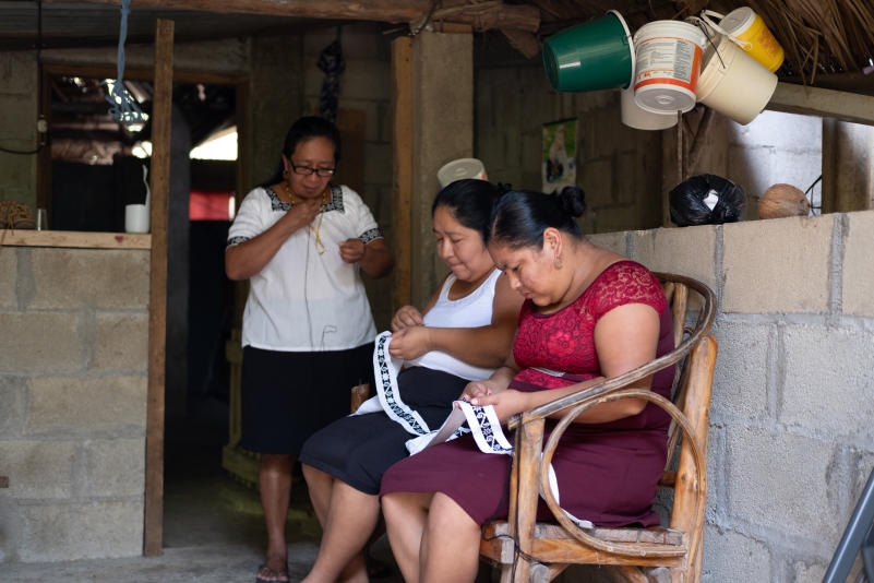 three Mayan ladies two seated sewing garments one standing looking on