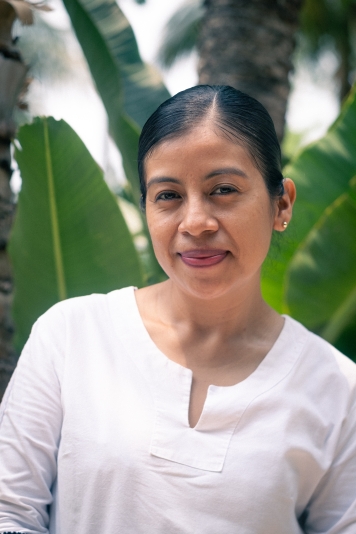 One Mayan woman in a white top standing in front of a large green leafed plant