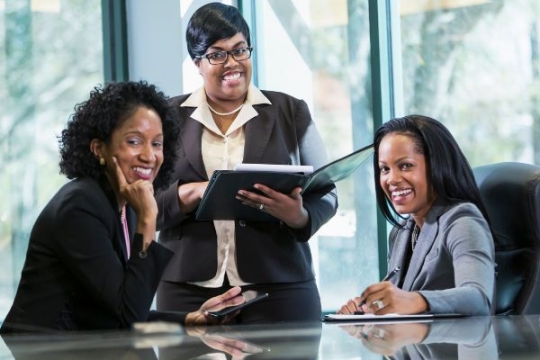 three black women in business attire in a boardroom. Two are seated and ne is standing the middle. They are smiling into the camera.