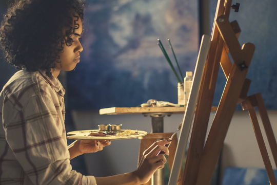 black female in white and brown plaid long-sleeved top sitting in front of an easel painting