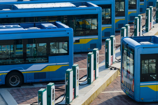 Row of blue electric buses parked at a charging station with multiple charging units on a paved lot.