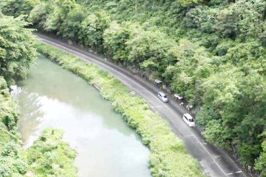 Aerial view of a winding road running alongside a calm river, surrounded by dense green forest and vegetation